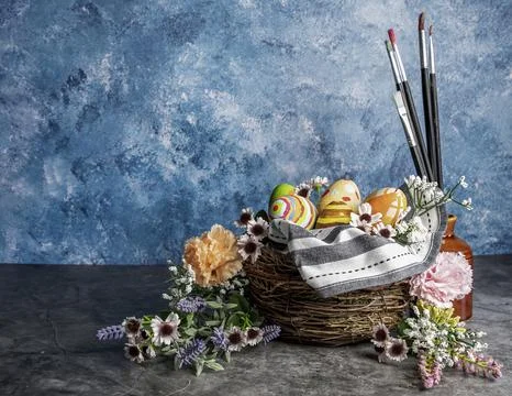 Paint brushes and colored Easter eggs in a straw corp with flowers Still life Stock Photos