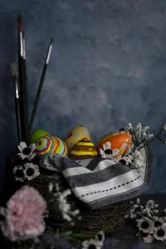 Paint brushes and colored Easter eggs in a straw corp with flowers Still life Stock Photos