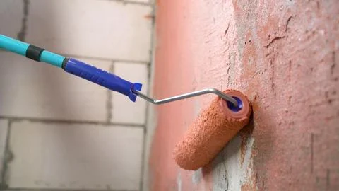 Paint roller applying red primer on rough concrete wall during renovation Stock Photos