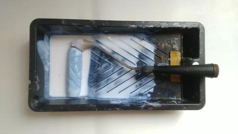 A paint roller rests in a black tray with white paint, brightly lit from abov Stock Photos