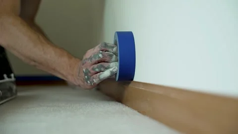 A painter carefully applies blue masking tape along the baseboard to prep clean Stock Footage 324802635
