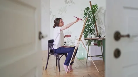 Painter in a workshop. View from the door. Young female artist draws sitting in Видео 156601344