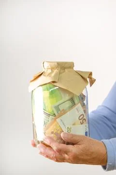 A pair of elderly hands hold a glass jar filled with paper money, including.. Stock Photos