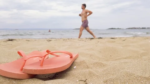 A pair of flip flops on a sandy beach. Man running along the beach. Stock Footage 114295475