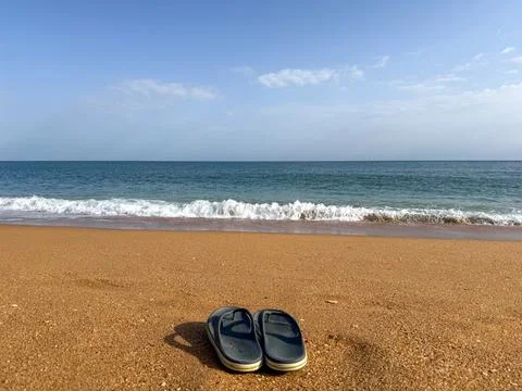 A pair of flip flops on a sandy beach with ocean waves and clear sky, summer  Stock-Fotos