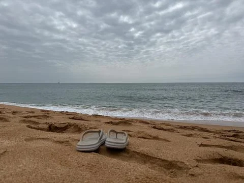 A pair of flipflops on a sandy beach with ocean waves and a cloudy sky, summe Stock Photos
