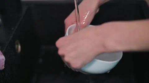 A pair of hands cleans a white bowl under running water in a kitchen sink. The Stock-Footage 337642824