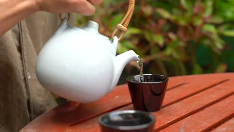 A pair of hands elegantly pouring tea from a white kettle into a small cup Stock Footage 234401016