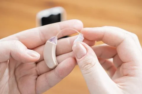 A pair of hands holds a beige behind-the-ear hearing aid to demonstrate its use Stock Photos
