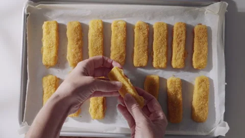 A pair of hands prepares breaded fish sticks on a parchment-lined baking tray, f Stock Footage 321870250