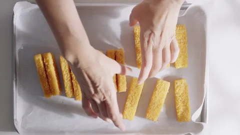 A pair of hands prepares breaded fish sticks on a parchment-lined baking tray.  Stock Footage 321872254