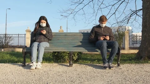 A pair in protective masks sit at opposite ends of a bench and use phones, Stockbeeldmateriaal 126247152