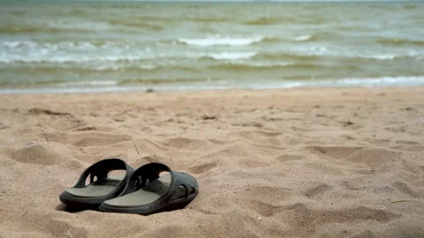 A pair of slippers lay on the beach with a small gentle lapping wave at Stock Footage 133313068