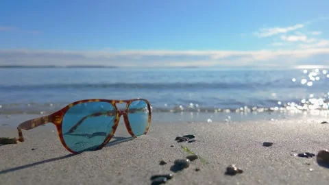 A pair of sunglasses resting on a sandy beach with gentle waves approaching Stock Footage 289065317