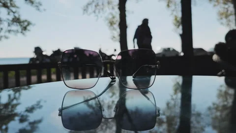 Pair of sunglasses sitting on a reflective table outdoors. In the blurred Video stock 347075970