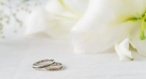 Pair of wedding rings on a white surface with beautiful white lilies flowers  Stock Photos