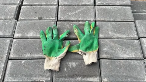 A pair of worn green work gloves with holes lies on gray paving stones 库存影片 341952034