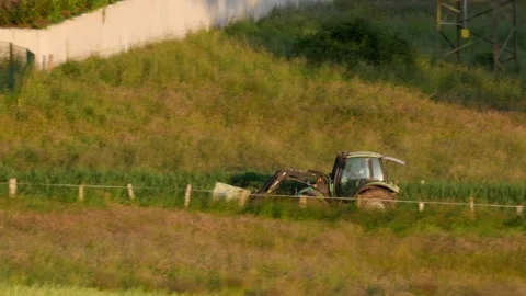 Pan: Farmer transporting hay bale on fork of tractor, driving on farm track Stock Footage 242940278