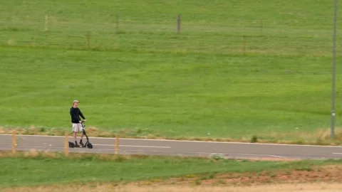 Pan: young man riding e-scooter on small rural road through pastures Stock Footage 246830886