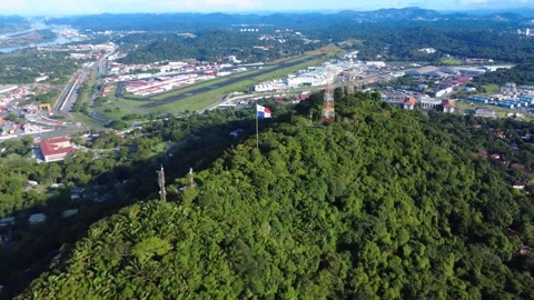 Panama flag flies high over lush green hill overlooking city and airport Stock Footage 323389393