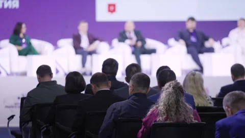 Panelists on stage with audience attentive delegates seated rows purple backdrop Stock Footage 328905157