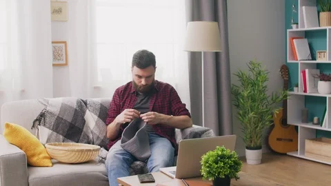 Panning of young man sitting on sofa and watching knitting tutorial on laptop Video stock 107351093
