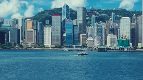 A panoramic view captures Hong Kong’s iconic skyline against lush green hills, 库存影片 338311010