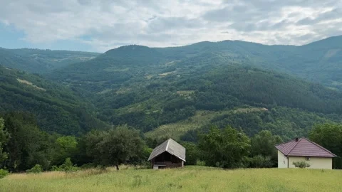 Panoramic view of forested mountains and a grassy valley with a rustic cabin in Stock Footage 344899587