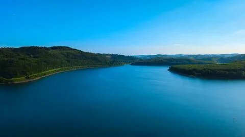 Panoramic view of lake surrounded by forested hills under blue sky. Stock Photos