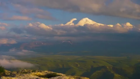 Panoramic view of Mount Elbrus peaks emerging from clouds over green hills in th Stock Footage 325812692