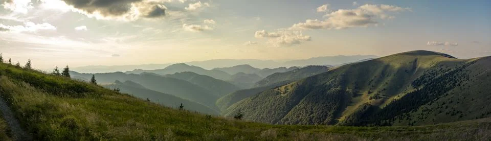 Panoramic view of sunlit rolling green hills and forested mountains under p.. Stock Photos