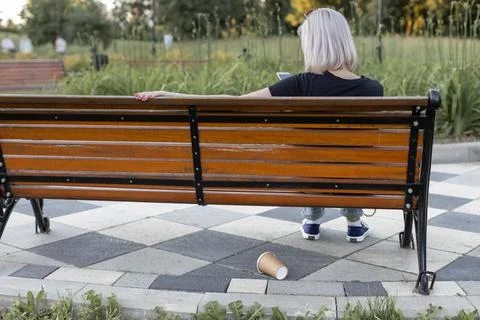 A paper cup is lying under a bench on which a women is sitting Stock Photos