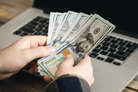Paper dollar bills in the hands of a man against the background of a laptop. Stock Photos