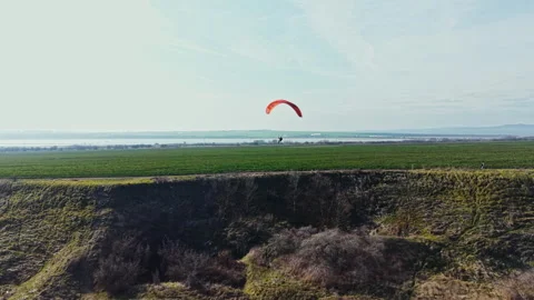 Paraglider flies above green fields near a body of water on a clear day with 스톡 동영상 336573563