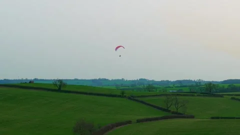 Paraglider gliding above rolling green countryside fields hedgerow farmland Stock Footage 334323628