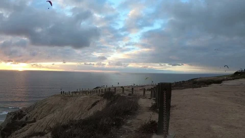 Paragliders fly past cliffs over ocean under a cloudy but colorful sky Stock Footage 82104323