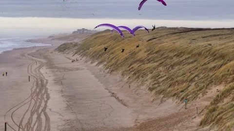 Paragliders soaring over grassy dunes along a misty coastline with cloudy skies Stock Footage 296804690