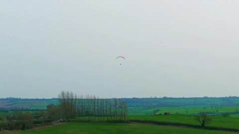Paramotor pilot gliding above green rural hills farmland hedgerows and tree Stock Footage 332218317