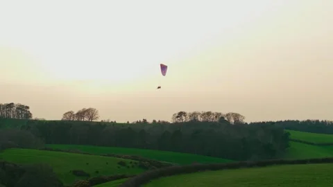 Paramotor pilot gliding above rolling green countryside hills and forest ridge Video stock 336541126