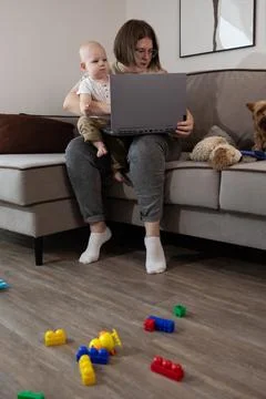 Parent on couch holding baby while using laptop with toys on floor Stock Photos