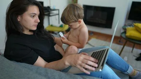 Parent multi tasking in front of laptop computer laying on couch. Toddler b.. Foto stock