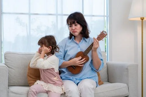Parent plays ukulele while child covers ears in living room during the day Stock Photos