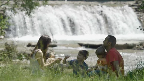 Parent with three children sitting on grass near flowing waterfall  Video stock 142034549