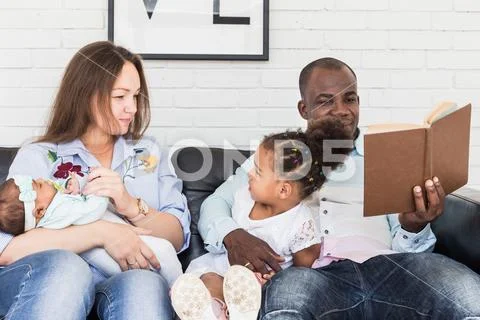 Parents read a book to children sitting on the couch. Happy multiethnic family Stock Photos