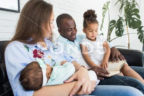 Parents read a book to children sitting on the couch. Happy multiethnic family Stock Photos