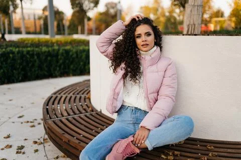 In the park on the bench bent under one leg sits a beautiful brunette in warm Stock Photos
