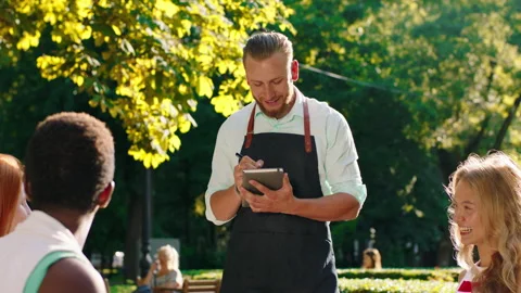 In a park with lots of trees, a very charming waiter with a beard is talking to Stock Footage 158832229
