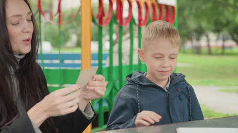 Park table white boy prepares school card, shy surprise for mom with colorful Stock Footage 333098582