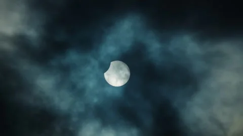 A partial solar eclipse shines brightly through thick, dark, and moody cloud Stock Footage 330455335
