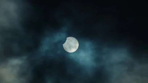 A partial solar eclipse shines brightly through thick, dark, and moody cloud Stock Footage 330455339
